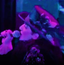 a blue-and-purple close-up of brian molko as malcolm singing into a microphone.